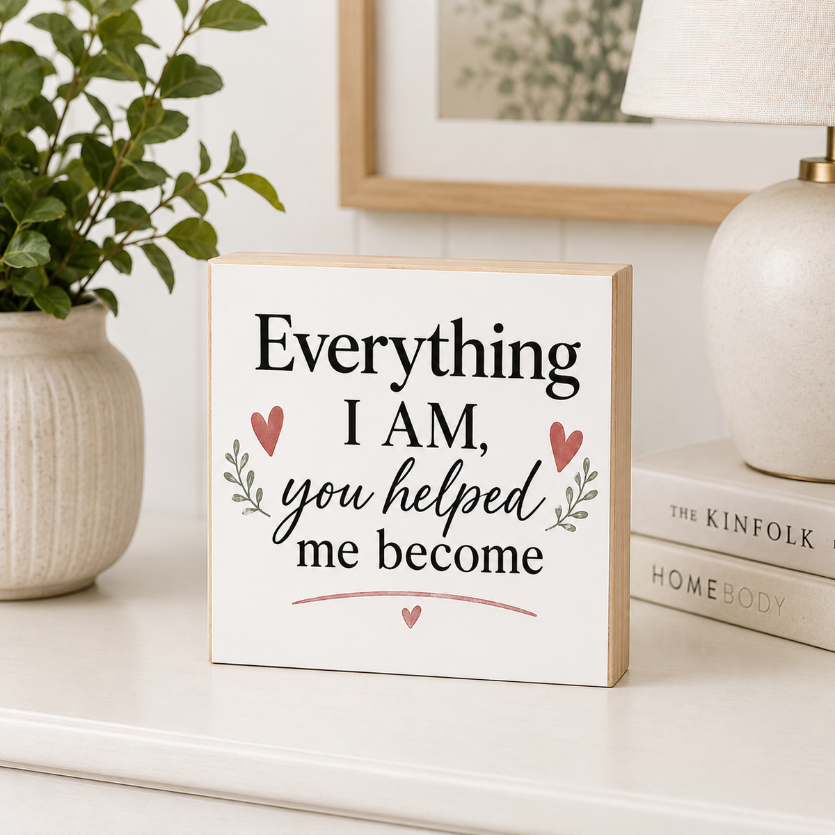 A square decorative wooden sign with a white face and natural wood edges, printed with the phrase 'Everything I AM, you helped me become' in black script and serif text, with small pink hearts and green leaf motifs. The sign is styled on a white shelf next to a potted plant, books, and a lamp.