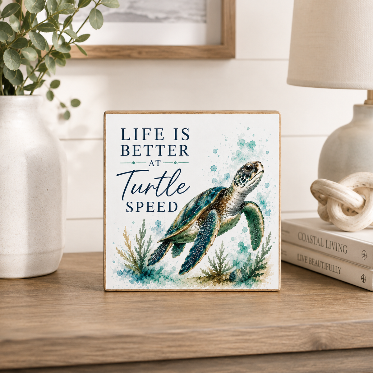A square decorative sign with a wooden border sits on a wooden surface in a styled home setting. The sign features a watercolor-style sea turtle and the text 'LIFE IS BETTER AT Turtle SPEED.
