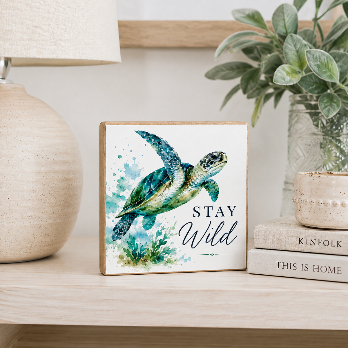 A square decorative wall sign with a watercolor sea turtle and the words “STAY Wild,” displayed on a light wood shelf beside a lamp, books, and a plant.