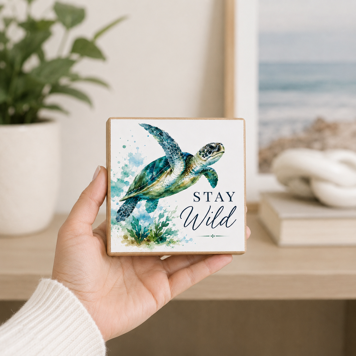 A hand holds a square wooden plaque featuring a watercolor sea turtle and the words 'STAY Wild' in navy script, set against a softly blurred home decor background with a plant and framed ocean print.
