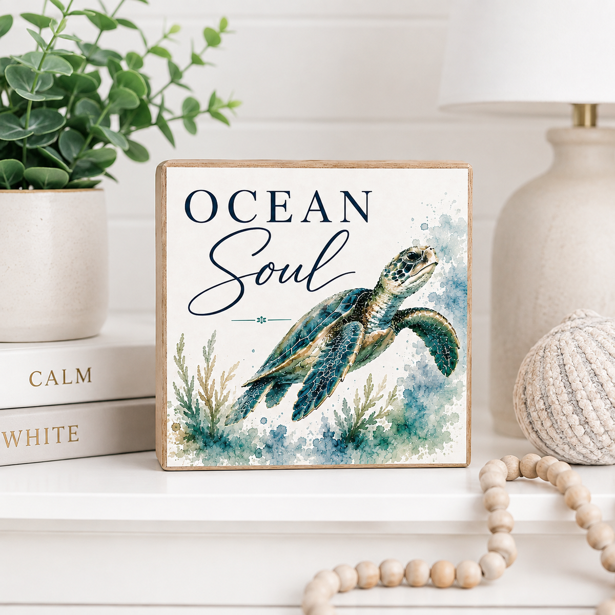 A square decorative wooden sign with a sea turtle illustration and the words “OCEAN Soul” in navy script, styled on a white shelf with a plant, books, a lamp, and a beaded necklace nearby.