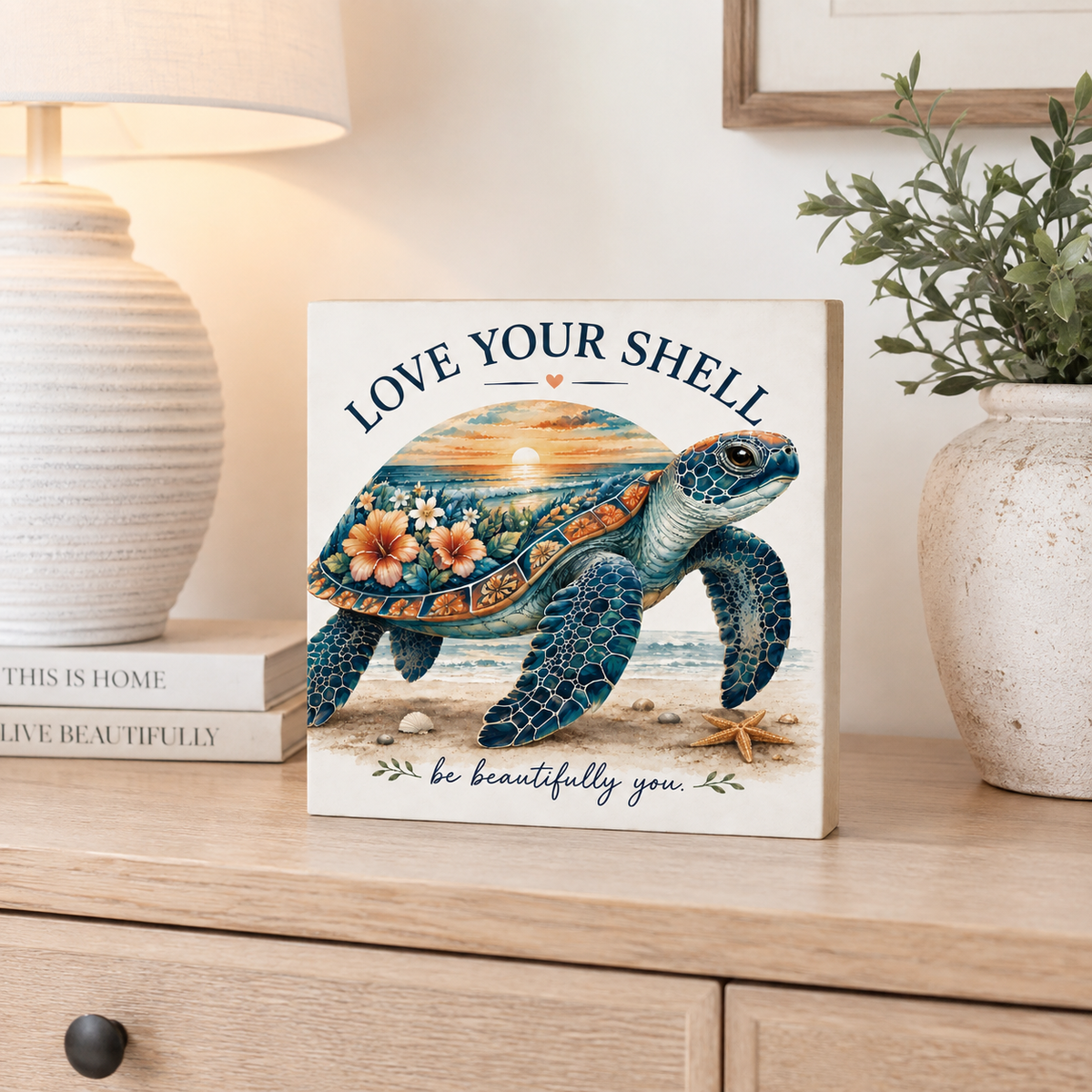 A square decorative sign featuring a colorful sea turtle illustration with the words 'LOVE YOUR SHELL' and 'be beautifully you.' displayed on a wooden console table in a styled home interior.