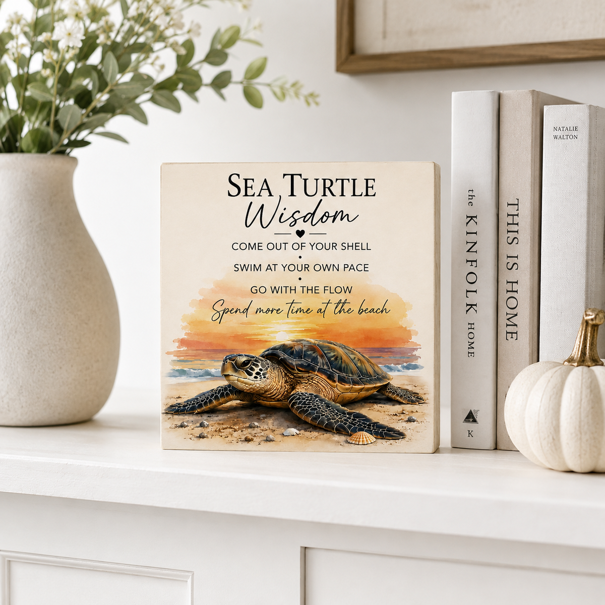 A square decorative sign featuring a sea turtle illustration and the text 'SEA TURTLE Wisdom' stands on a white mantel beside a vase with greenery, books, and a small pumpkin.