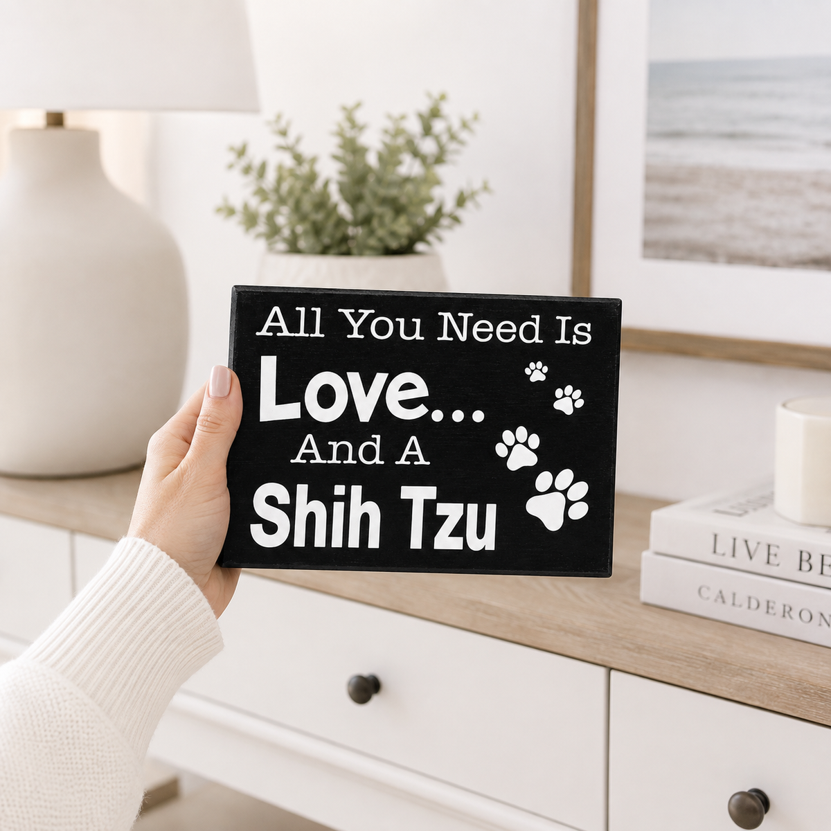 A hand in a white ribbed sweater holds a black rectangular sign that reads 'All You Need Is Love.