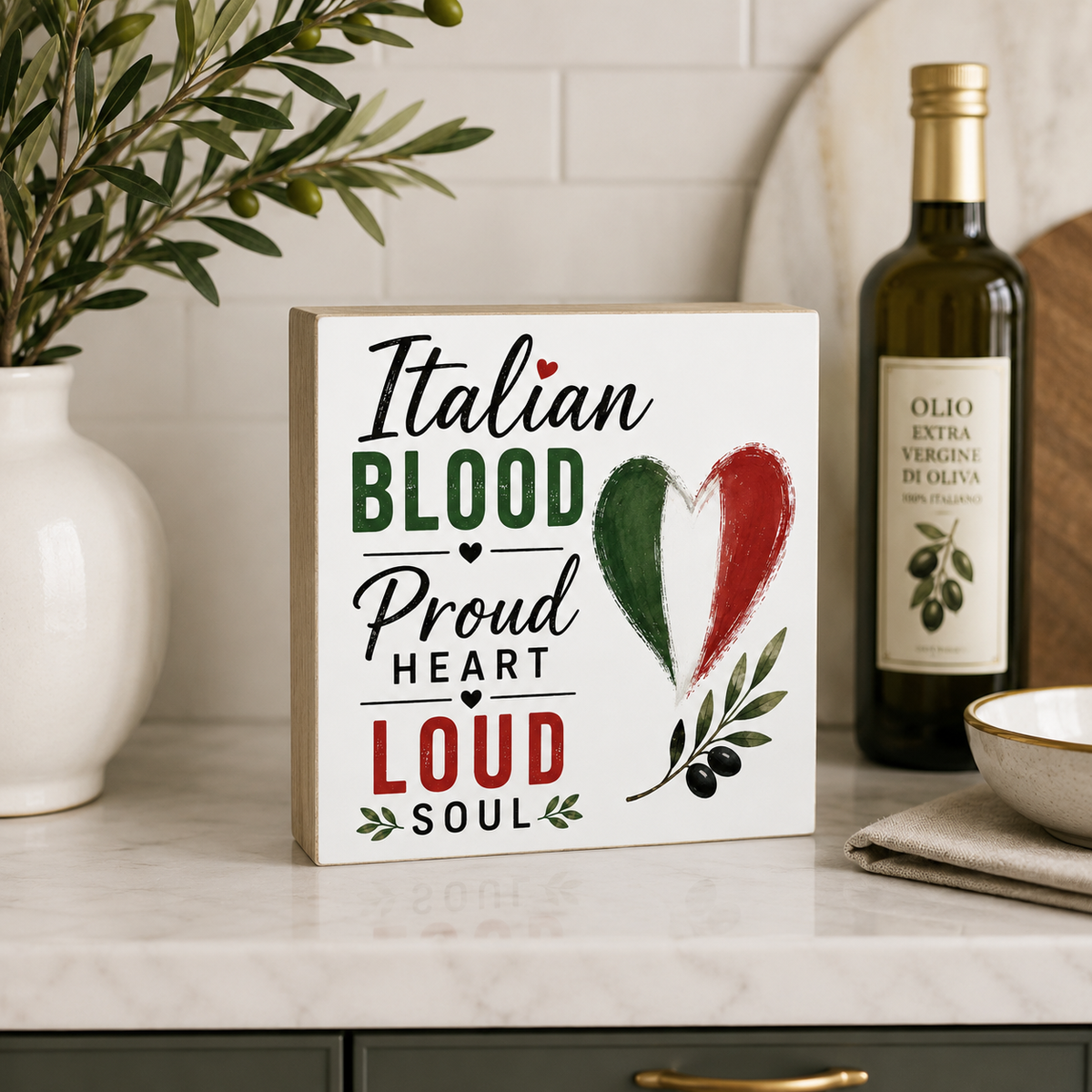 A square white decorative sign with the phrase 'Italian Blood Proud Heart Loud Soul' in black and green lettering, plus a heart painted in green, white, and red and olive branch illustrations. The sign sits on a marble countertop in a kitchen setting, with a bottle of olive oil, a white vase with greenery, and a bowl nearby.