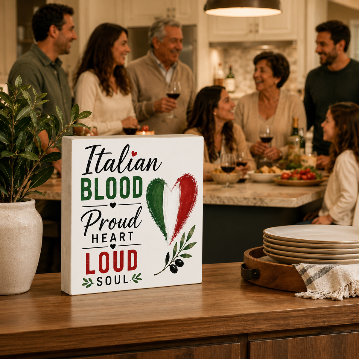 A square white decorative sign on a wooden kitchen counter reads “Italian Blood Proud Heart Loud Soul” with a green, white, and red heart and olive branch illustration. In the background, a group of people gather around a kitchen island with wine glasses and food.