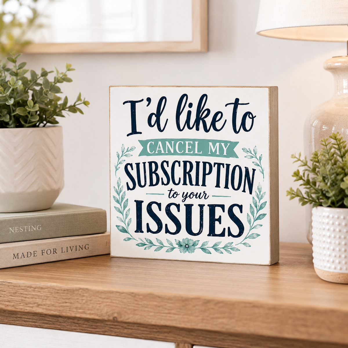 A square decorative sign with the text 'I'd like to CANCEL MY SUBSCRIPTION to your ISSUES' in navy and teal lettering, styled on a wooden table with books, potted plants, and a lamp in a home setting.