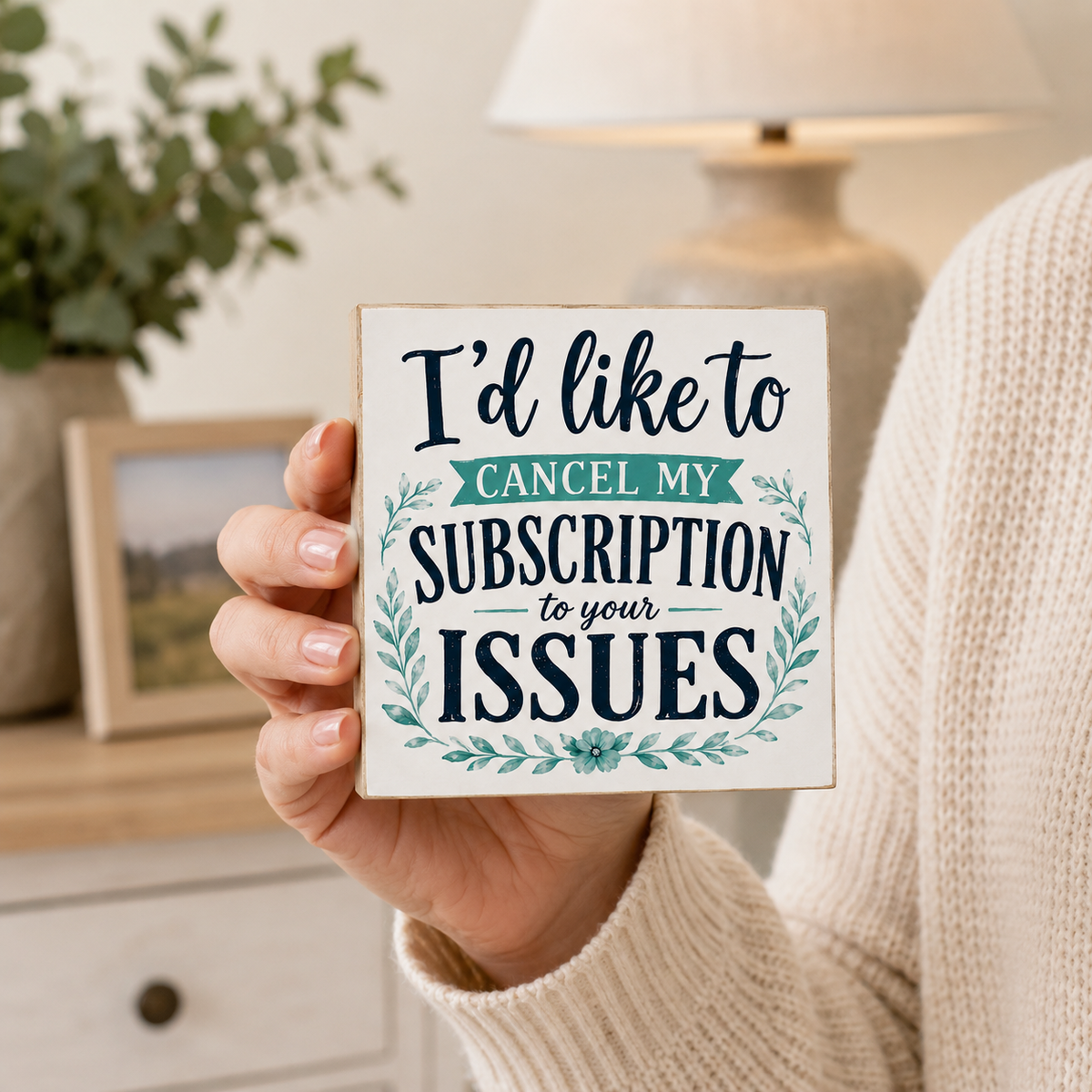 A person in a cream knit sweater holds a square white decorative sign with navy and teal text that reads, 'I'd like to cancel my SUBSCRIPTION to your ISSUES.' The background shows a softly blurred indoor room with a lamp, framed picture, and greenery.