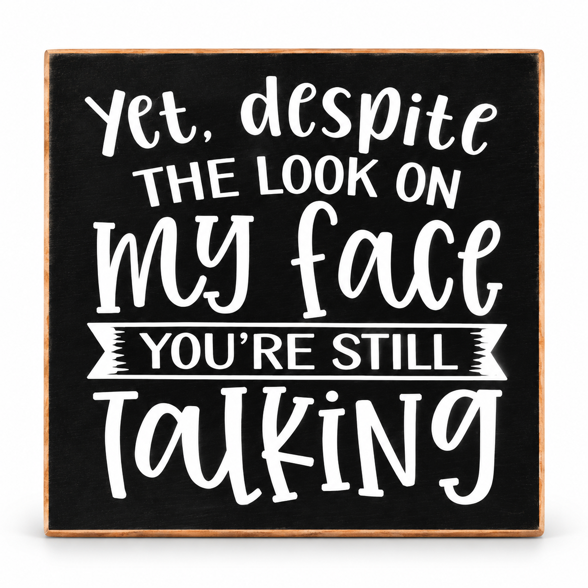 A square decorative sign with a black chalkboard-style face, a light wood border, and white hand-lettered text that reads, “Yet, despite the look on my face you’re still talking.”.