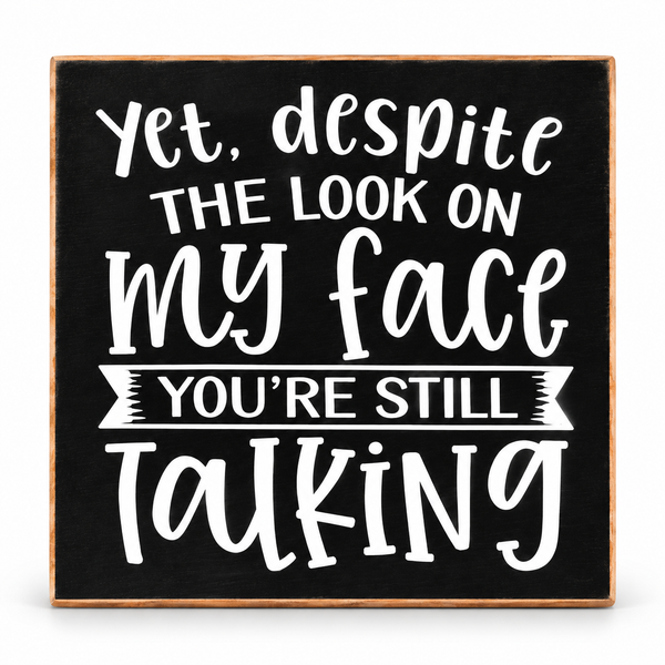 A square decorative sign with a black chalkboard-style face, a light wood border, and white hand-lettered text that reads, “Yet, despite the look on my face you’re still talking.”.
