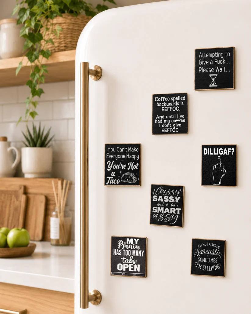 A white refrigerator in a kitchen setting with several black square magnets featuring humorous sayings in white script and block lettering. The magnets are arranged on the door next to a brass handle.