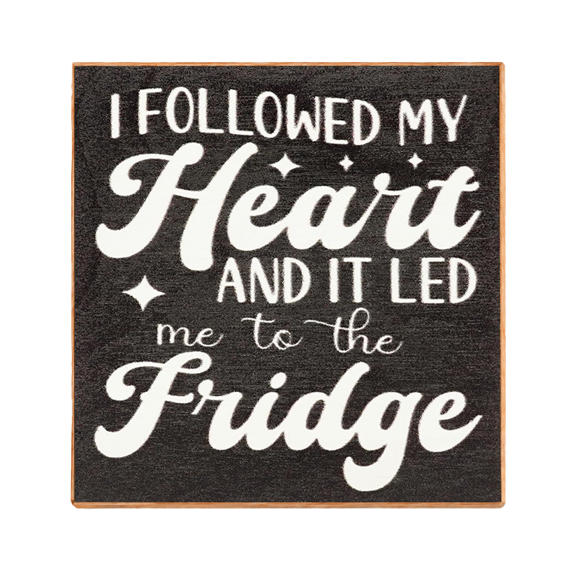 A square decorative sign with a dark charcoal textured background and a light wood border. White lettering reads, 'I FOLLOWED MY Heart AND IT LED me to the Fridge' with small star accents.