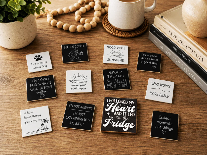 A styled flat-lay of multiple small square metal nameplates or magnets with humorous quotes, arranged on a wooden tabletop with a mug, books, beads, and a plant nearby.