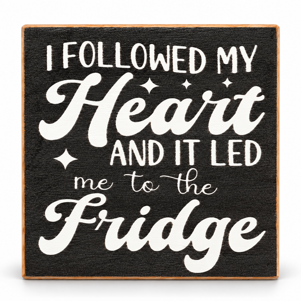 A square decorative sign with a dark charcoal-textured background and a light wood border. White lettering reads, 'I FOLLOWED MY Heart AND IT LED me to the Fridge' with small star accents.