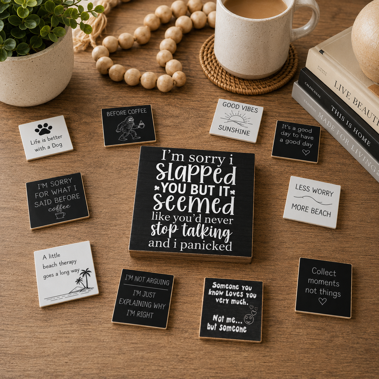 A styled flat-lay of multiple small square sign plaques with humorous and self-care quotes on a wooden tabletop, surrounded by a mug, books, beads, and a plant.