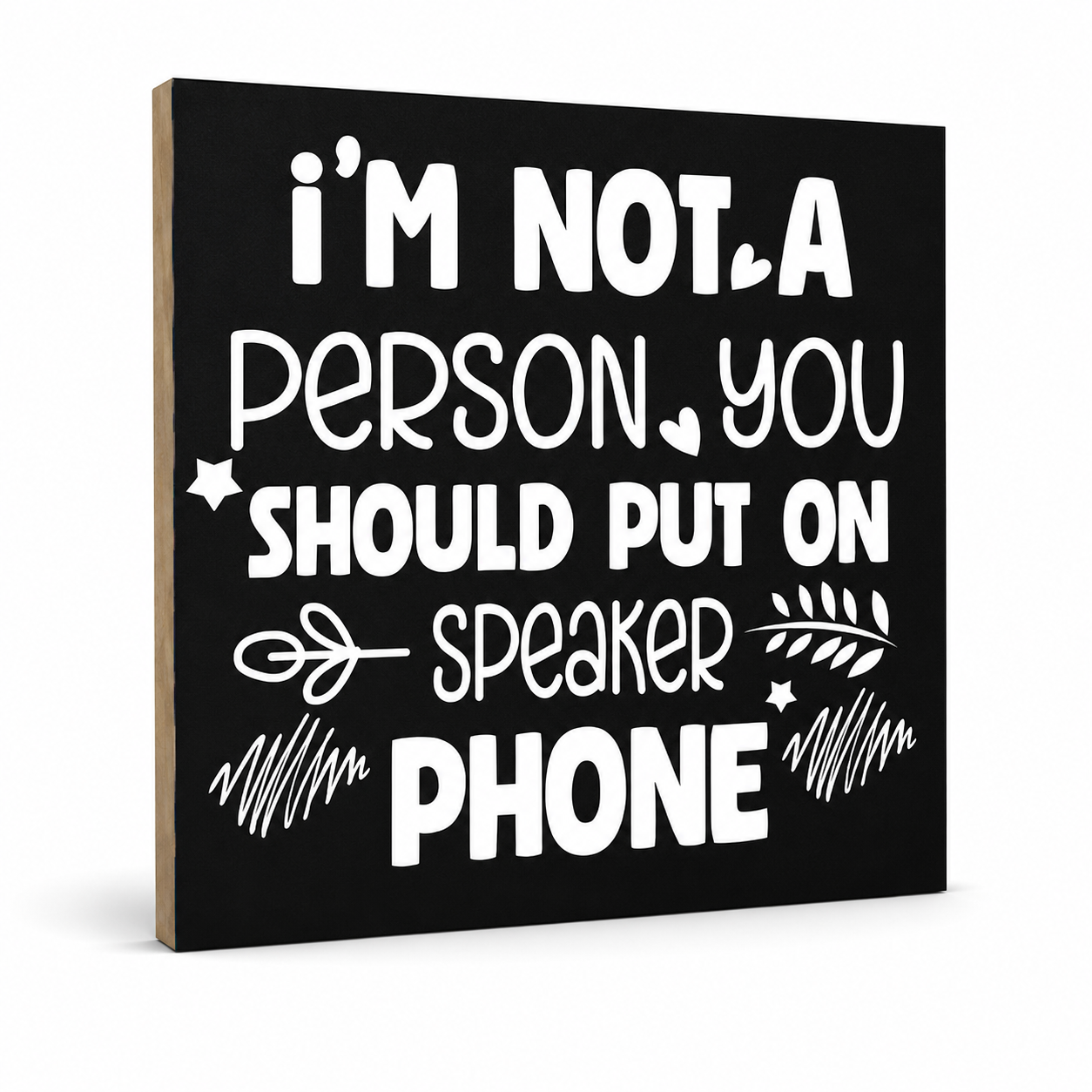 A square black decorative sign with a light wood edge, featuring white text that reads, 'i'M NOT A PERSON YOU SHOULD PUT ON SPEAKER PHONE' with small hand-drawn graphic accents, shown on a white background.