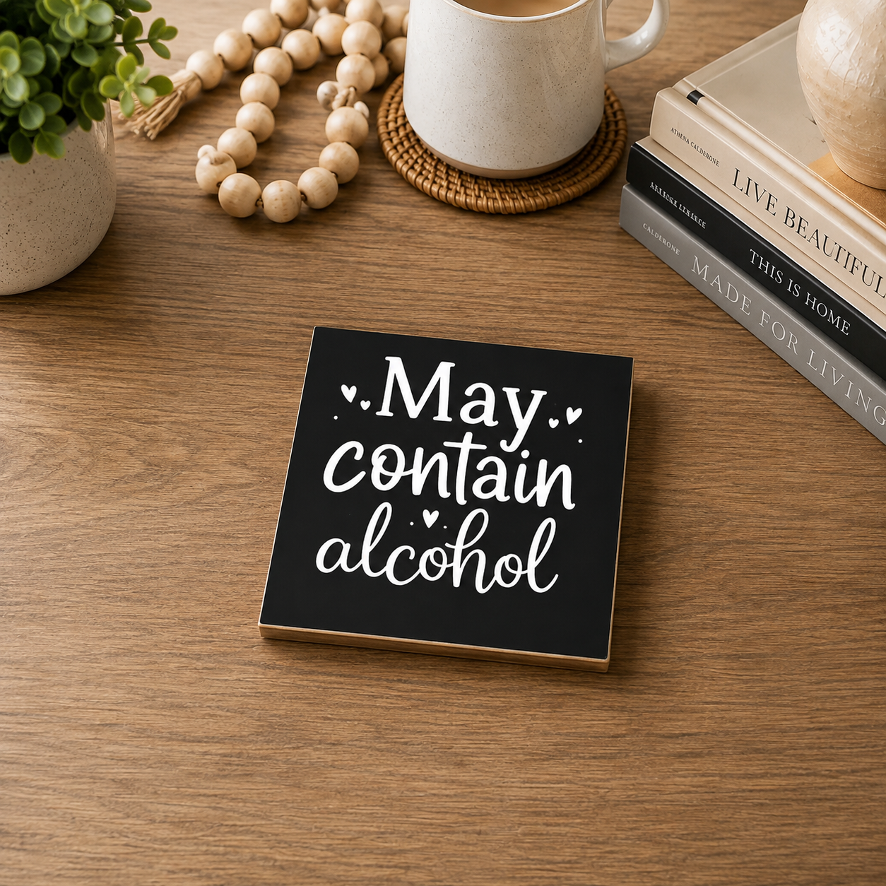 A square black decorative sign with white text reading 'May contain alcohol' sits on a wooden tabletop, surrounded by a mug, books, a plant, and a bead garland.