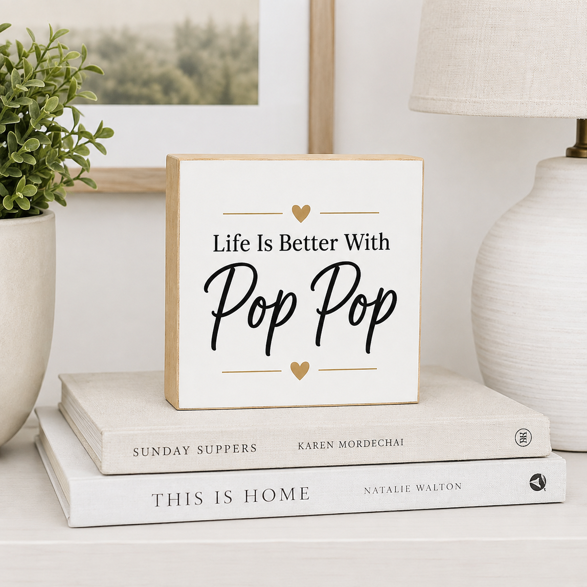 A square decorative wooden sign with the phrase 'Life Is Better With Pop Pop' in black script and serif text, accented with small gold hearts and lines. The sign is styled on top of two books on a white surface, with a potted plant and a lamp in the background.