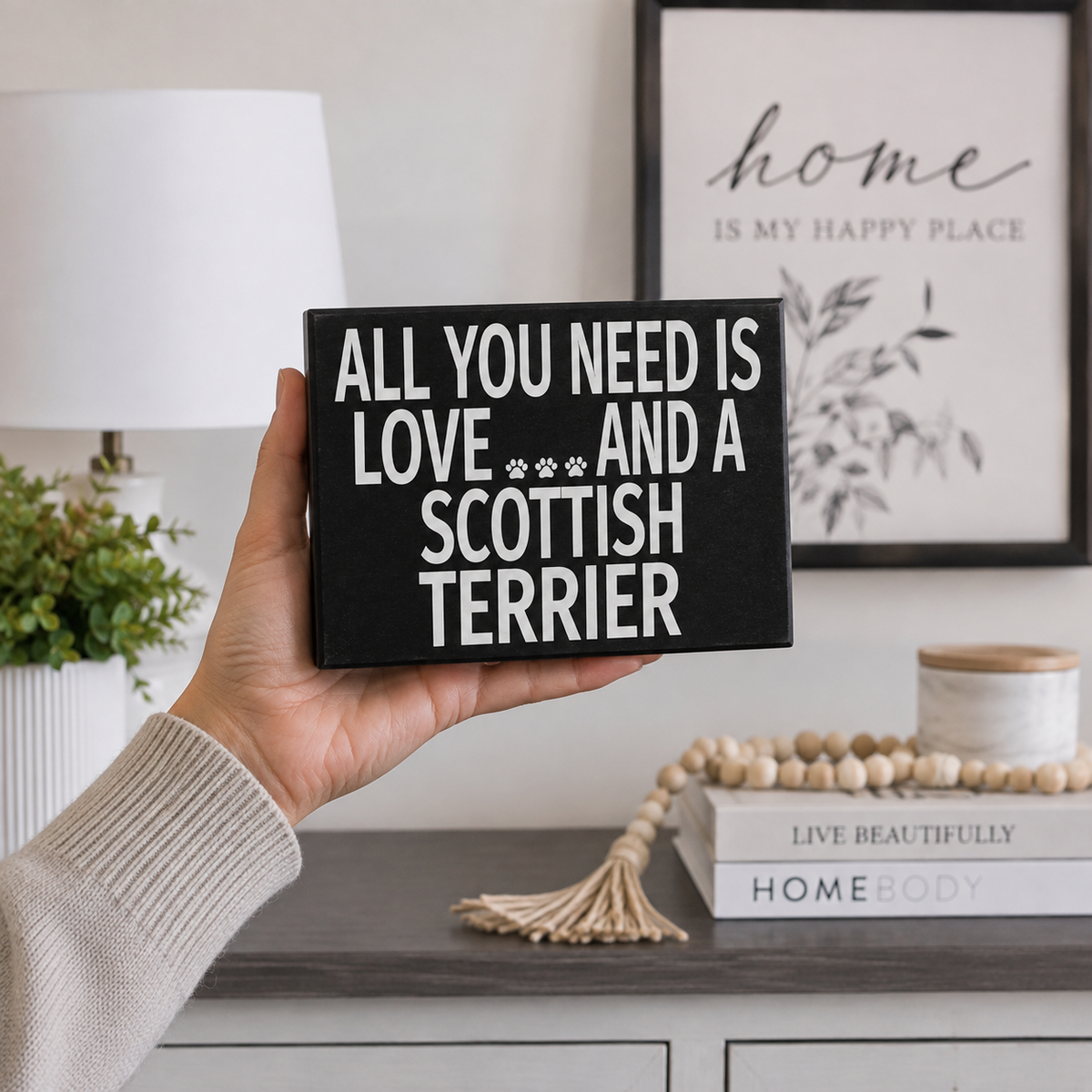 A hand holds a black rectangular sign with white text that reads 'ALL YOU NEED IS LOVE *** AND A SCOTTISH TERRIER' in a styled home interior setting.