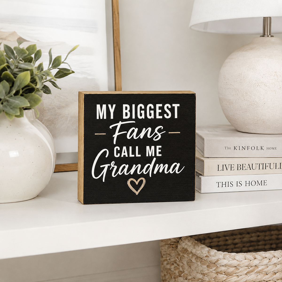 A rectangular black wooden sign with natural wood edges reads “MY BIGGEST Fans CALL ME Grandma” in white lettering, with a small gold heart outline near the bottom. It sits on a white shelf in a styled home decor setting with a vase, framed art, books, and a lamp.