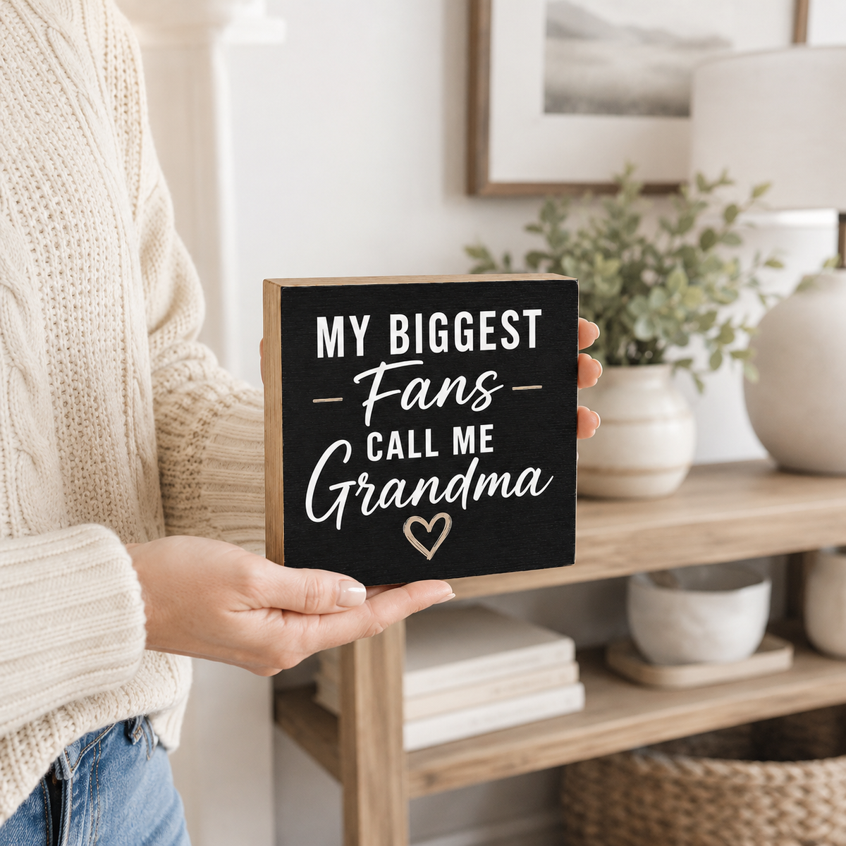 A person in a cream knit sweater holds a square black sign with the phrase 'MY BIGGEST Fans CALL ME Grandma' in white lettering and a small heart at the bottom. The scene is set in a bright, modern living room with a wooden shelf, potted plants, and a lamp in the background.