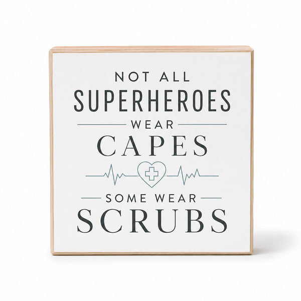 A square decorative wooden sign with a light natural wood frame and a white face, printed with the text 'NOT ALL SUPERHEROES WEAR CAPES SOME WEAR SCRUBS' in dark gray lettering with a light blue heart monitor line and medical cross symbol in the center, shown on a white background.