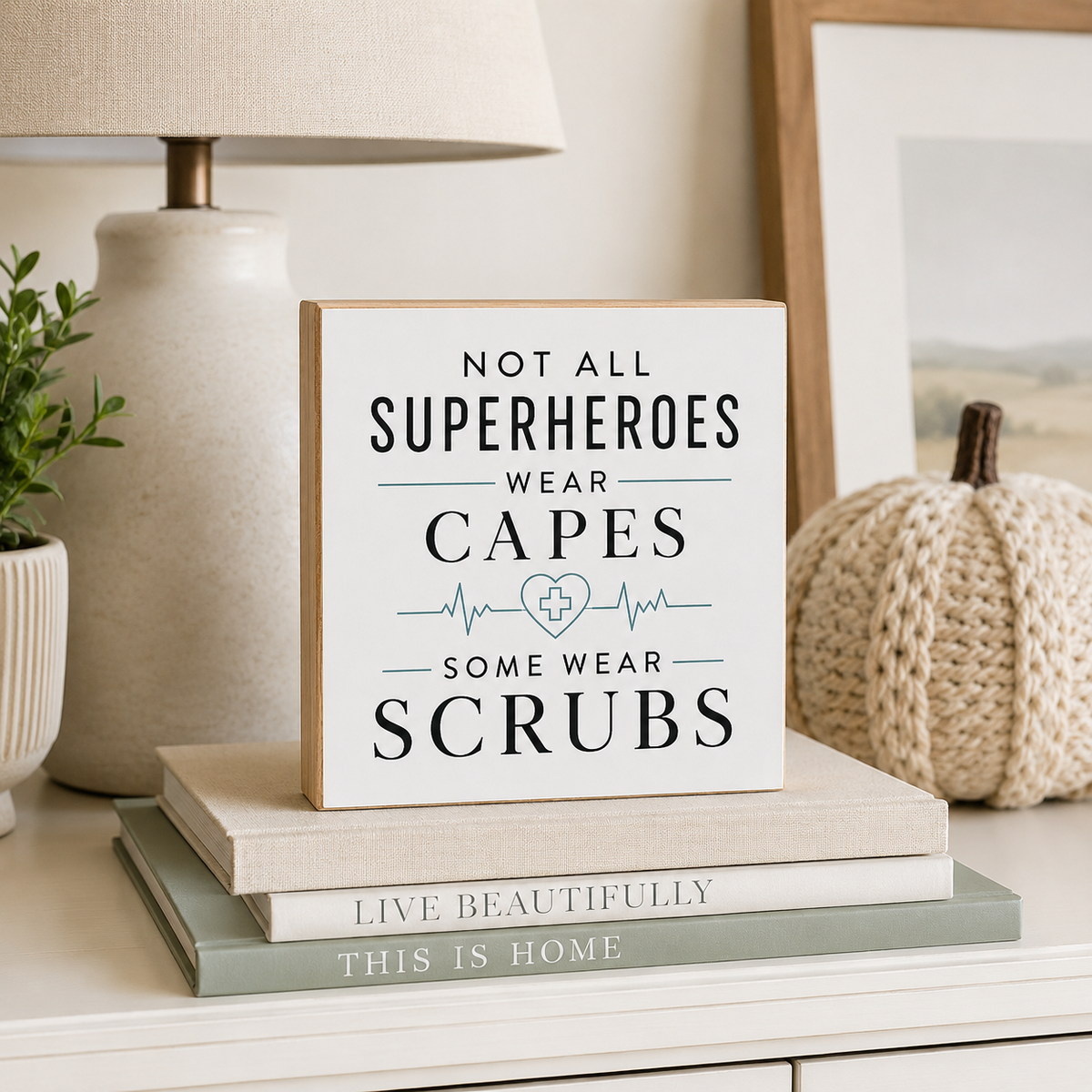 A square white decorative sign with the phrase 'NOT ALL SUPERHEROES WEAR CAPES SOME WEAR SCRUBS' in dark navy text and a small blue medical cross/heartbeat graphic, displayed on stacked books on a tabletop in a styled home setting.