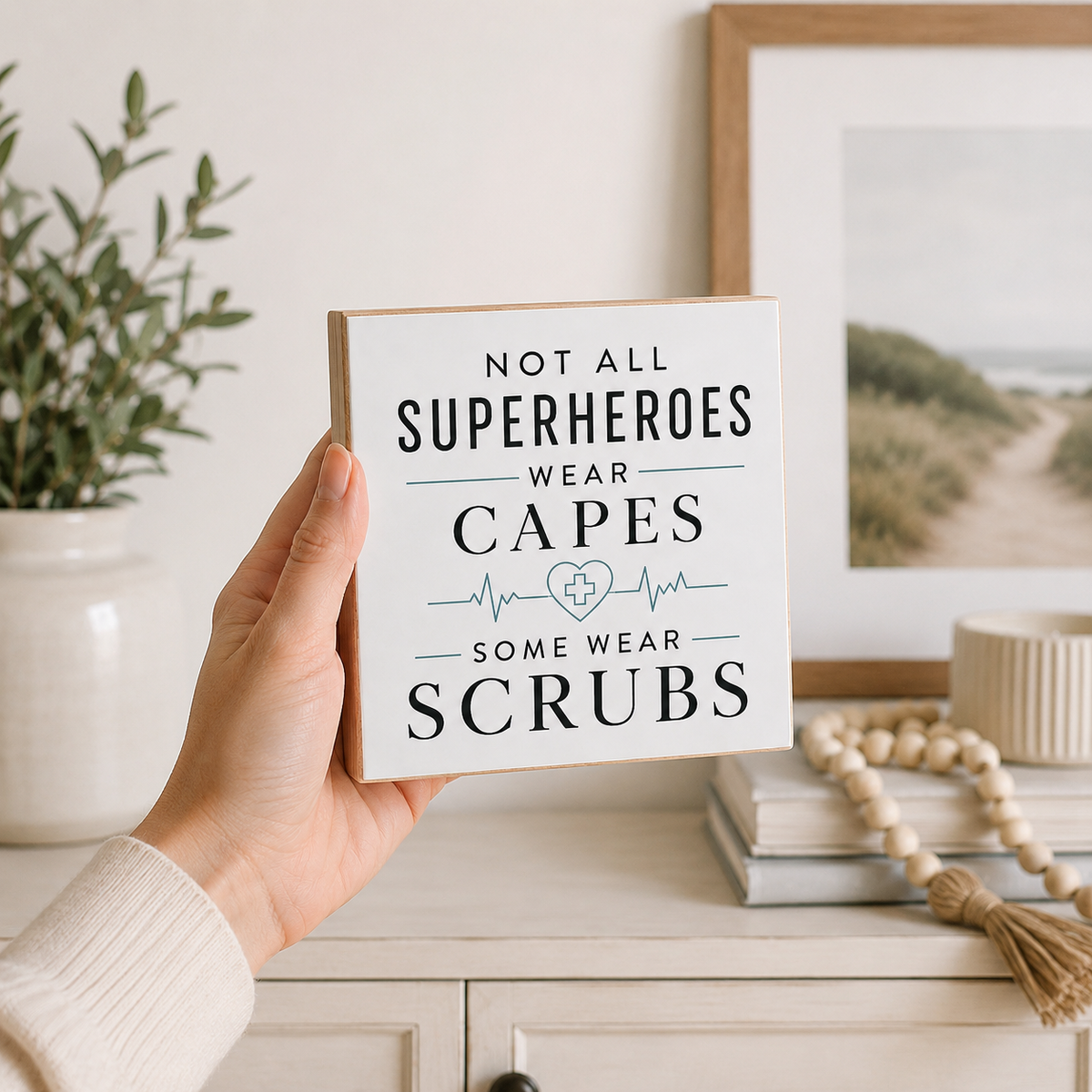 A hand holds a square white decorative sign with the text 'NOT ALL SUPERHEROES WEAR CAPES SOME WEAR SCRUBS' in dark lettering, styled in a bright, modern room setting with a framed print, books, and a plant in the background.