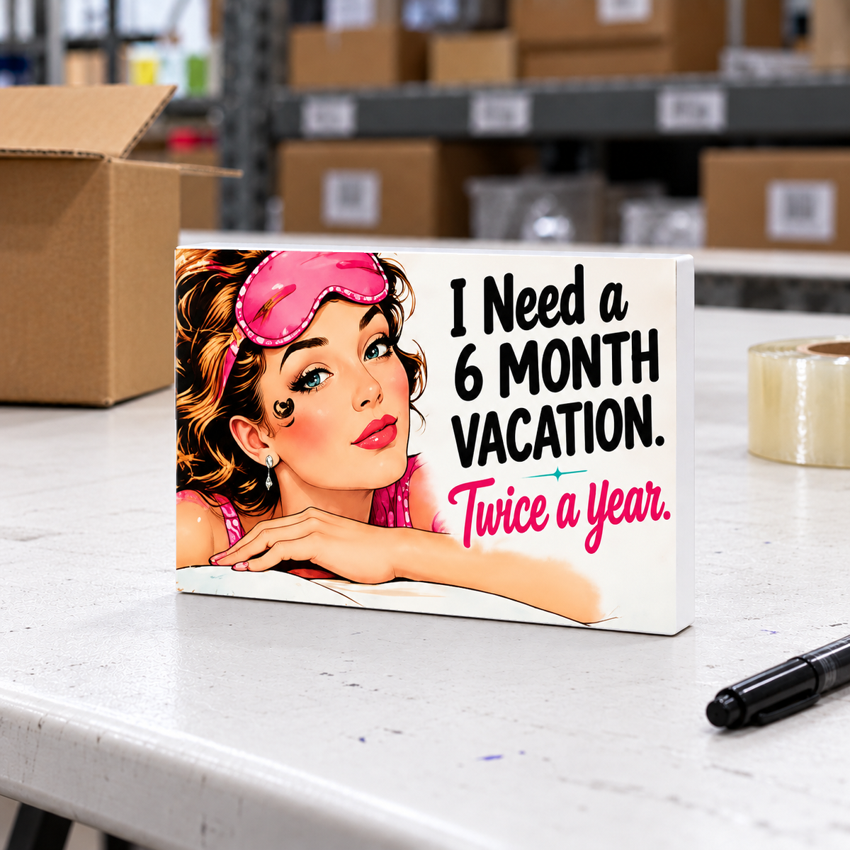 I Need a 6 Month Vacation Twice a Year mini sign on a white folding warehouse table with packing supplies.