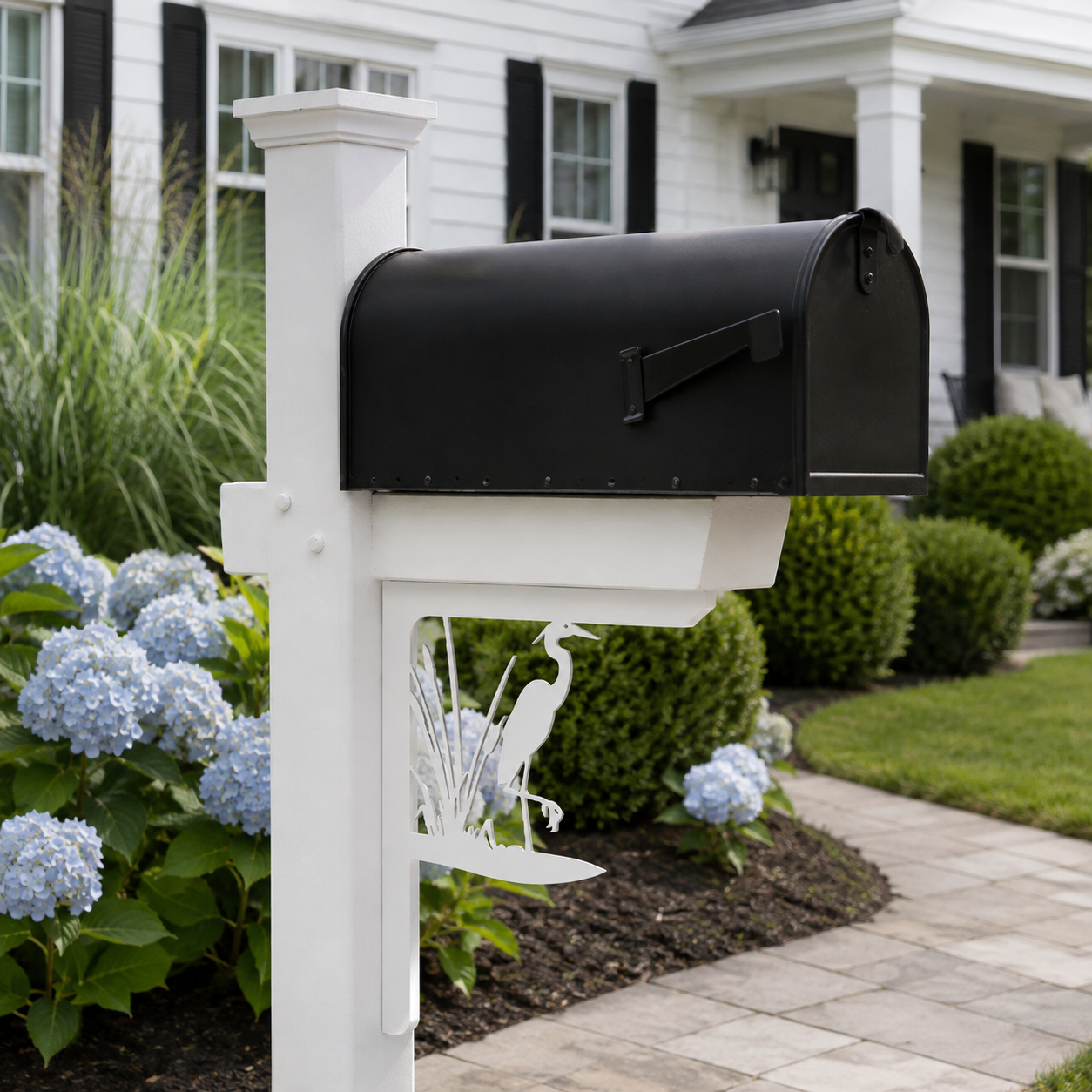 A black metal mailbox mounted on a white post in a landscaped garden in front of a white house with black shutters. A white metal heron decoration is attached below the mailbox.