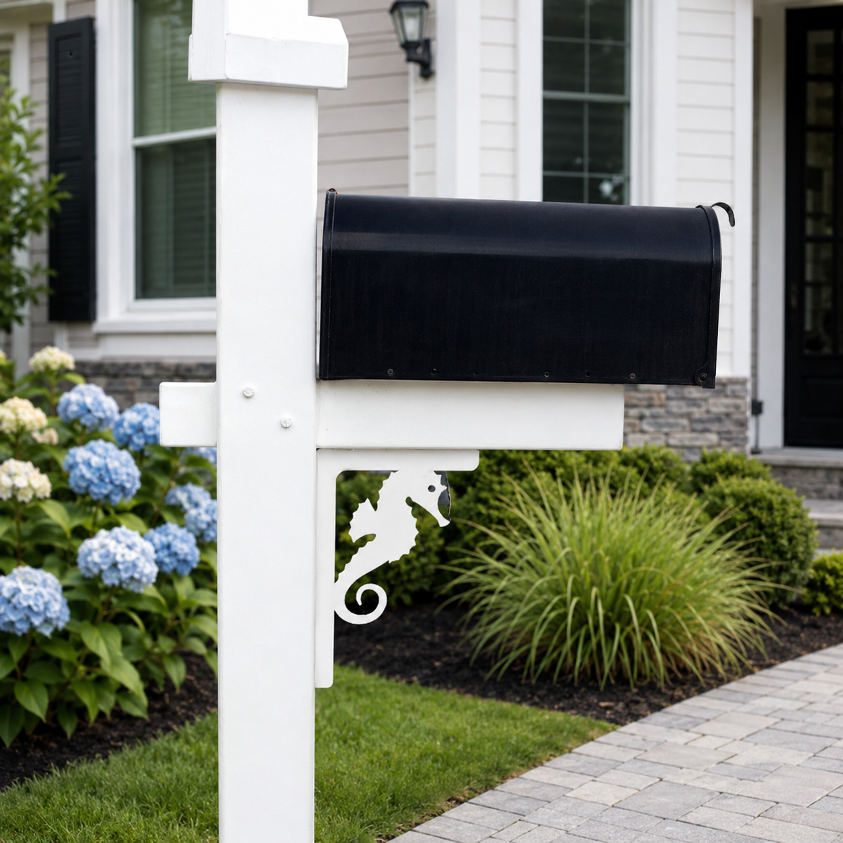 A black mailbox mounted on a white post with a decorative white seahorse accent, set in a landscaped garden in front of a house.