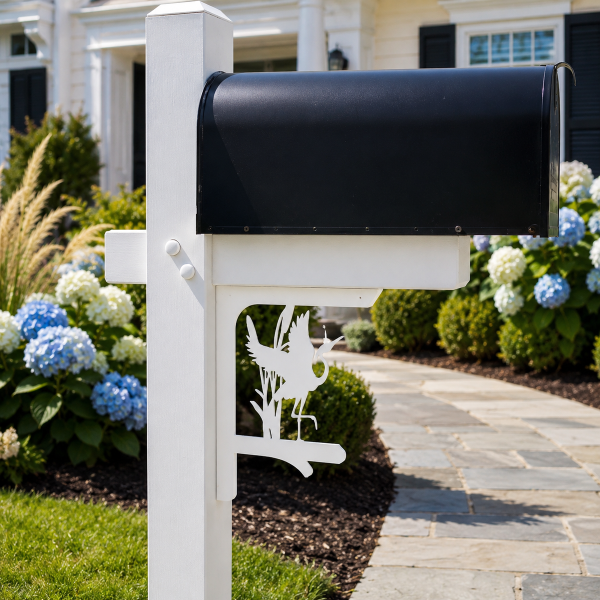 A white mailbox post with a black mailbox and a white metal bird silhouette cutout, set in a landscaped garden with hydrangeas and a stone walkway.