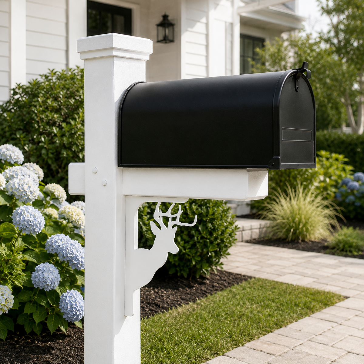 A black metal mailbox mounted on a white post with a decorative deer head cutout, set in a landscaped front yard with green shrubs, blue hydrangeas, and a paved walkway.