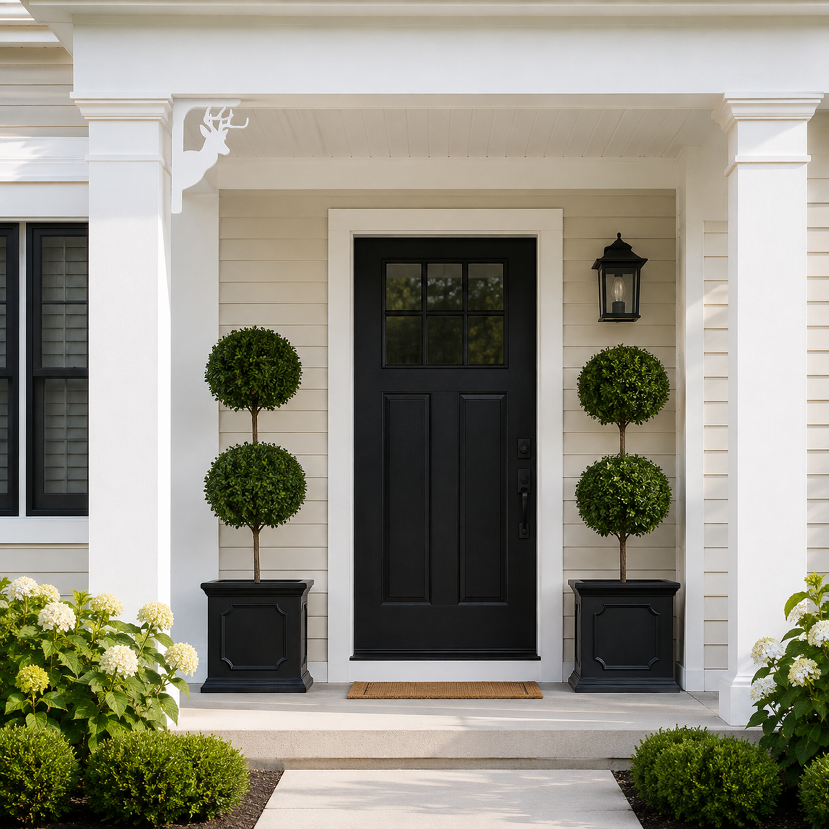 A front porch entryway with a black paneled door centered between white columns, flanked by two black planters holding rounded green topiary trees. The house siding is light beige, with a black wall lantern and landscaped shrubs at the base.
