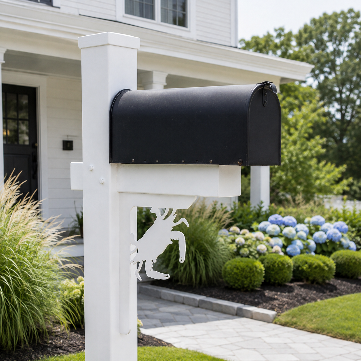 A black metal mailbox mounted on a white post in a landscaped front yard with a house, shrubs, and a stone walkway visible in the background.